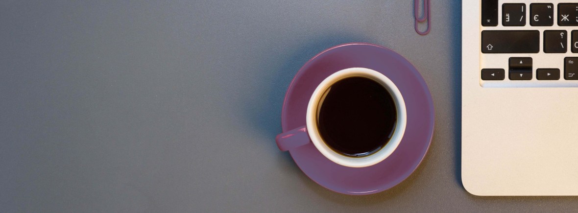 A partial laptop, purple pencil, purple paperclip, and purple coffee cup sitting on a grey desk.