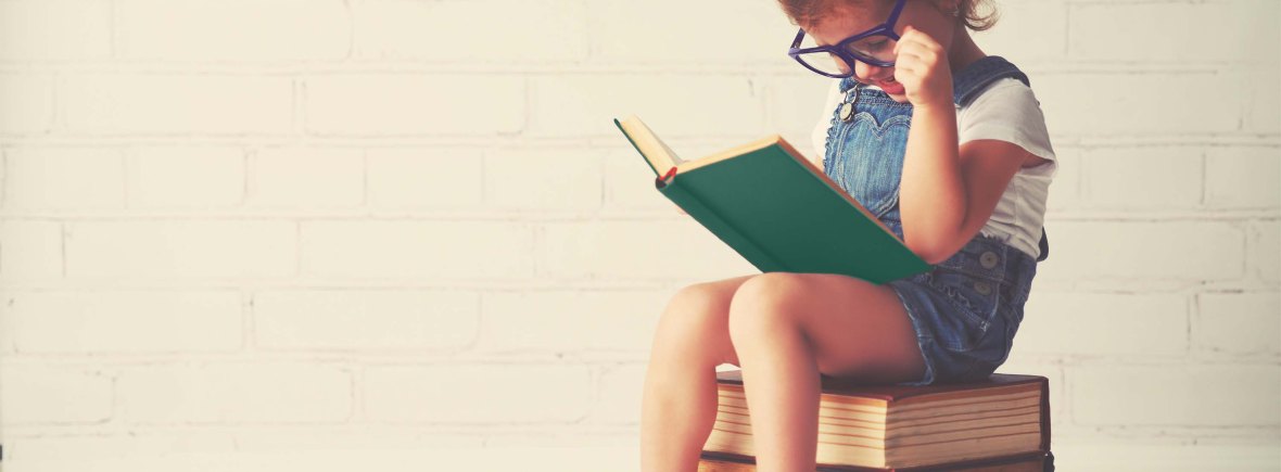 A young child sitting on a small pile of books reading a book. The child wears purple glasses and jean overalls.