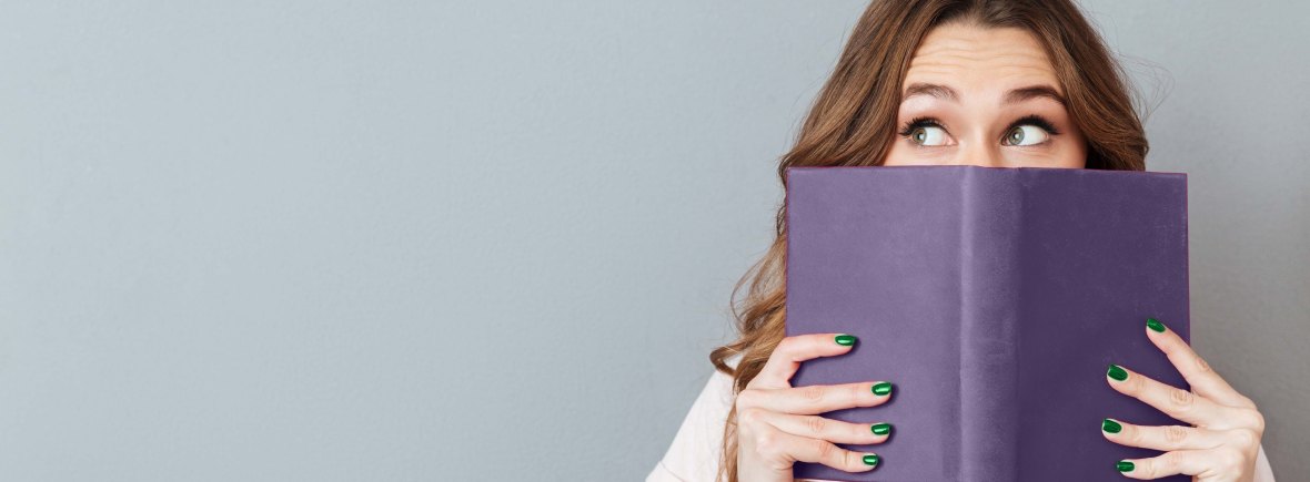 A young woman with brown hair hides behind a purple book. Her eyes are the only thing visible above the book.